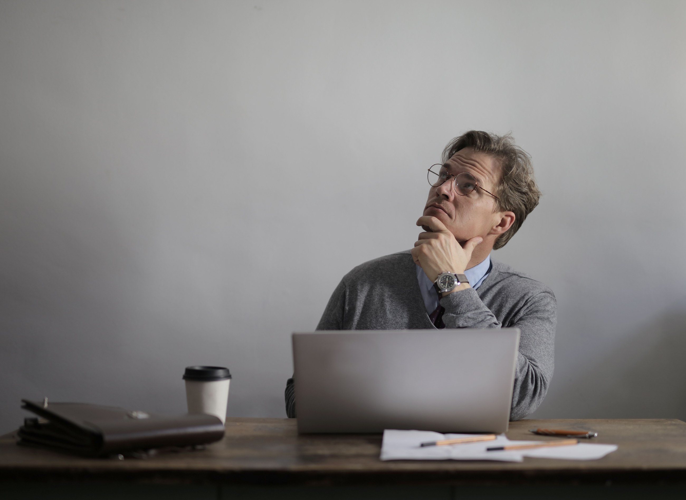 Hombre con lentes en actitud reflexiva, con la mano en la barbilla, frente a una laptop en un escritorio con café y documentos